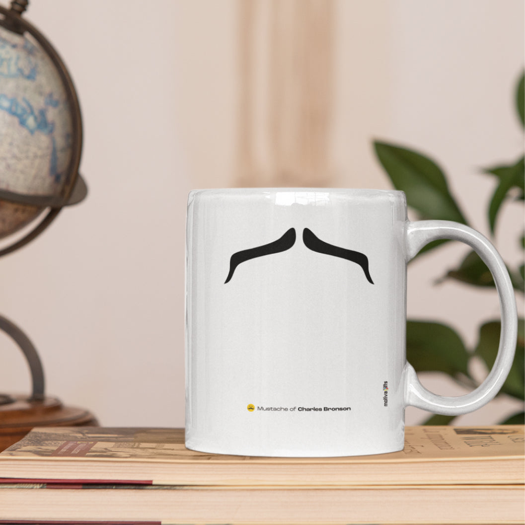 White ceramic mug with a drawing of Charles Bronson’s mustache featuring on a set of book. 