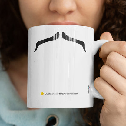 Woman holding a white ceramic mug with a drawing of Charles Bronson’s mustache, set against a plain white background.