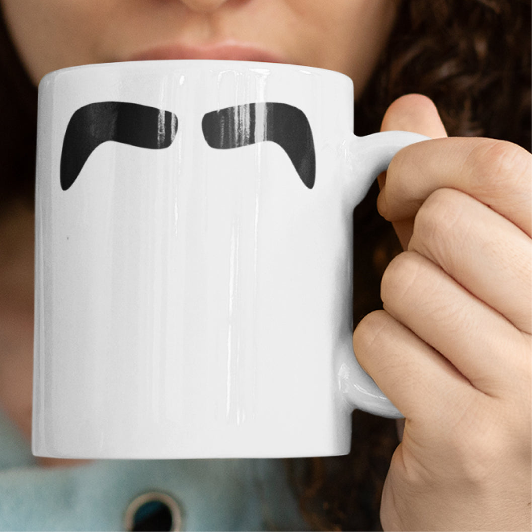 Woman holding a white ceramic mug with a drawing of John Lennon’s mustache, set against a plain white background.